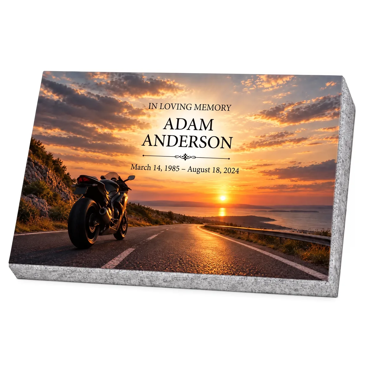 Memorial plaque in glass with a motorcycle on a coastal road at sunset, on a granite base