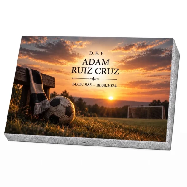Placa conmemorativa de vidrio con balón de fútbol, bufanda y portería al atardecer, sobre base de granito